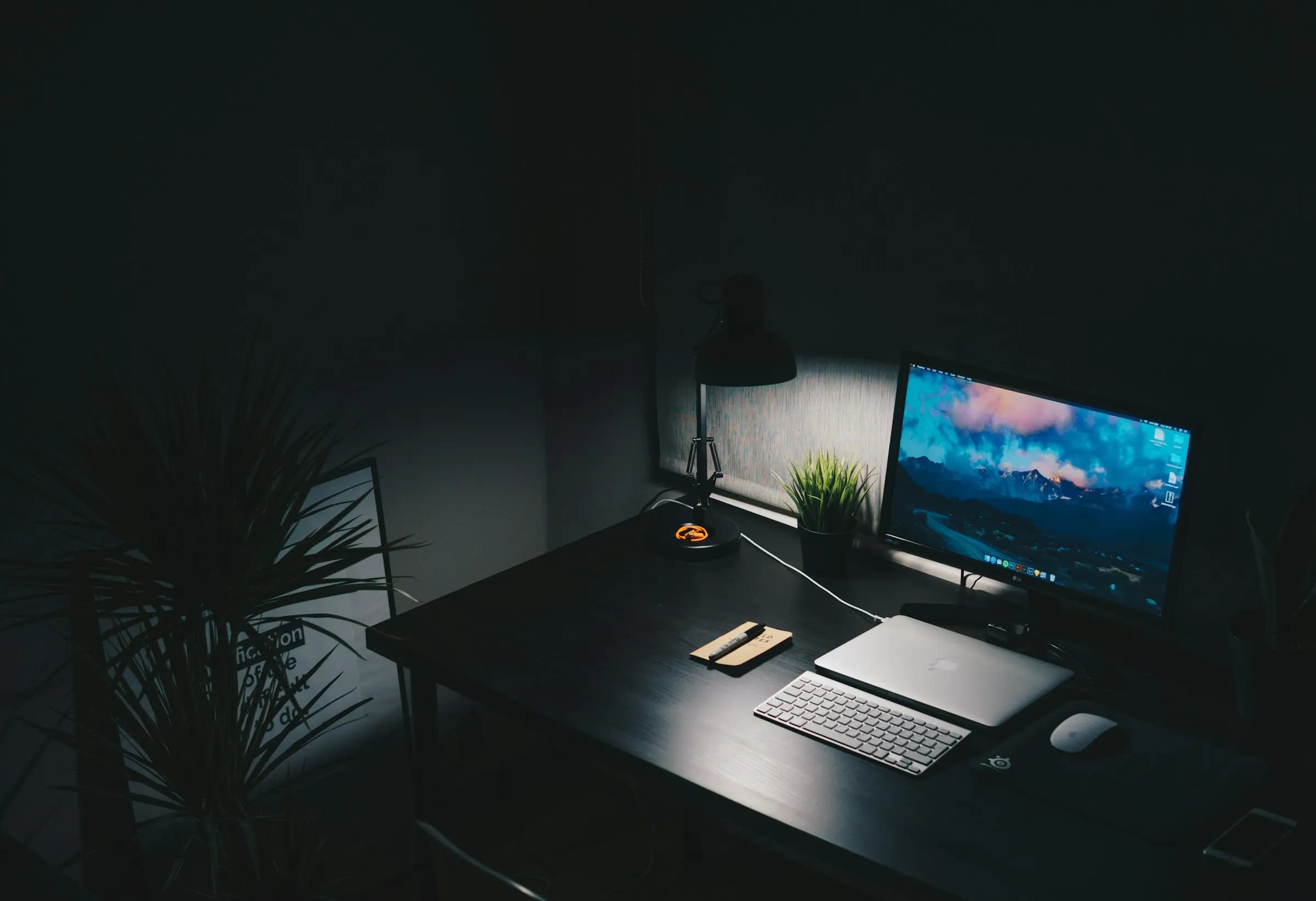 A dark studio desk at night, a monitor glowing beside a desk lamp